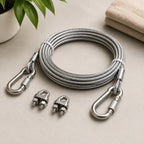 A coiled silver steel cable with two metal carabiner clips and two matching connector hardware pieces, arranged on a light beige surface with a plant leaf and folded fabric in the background.
